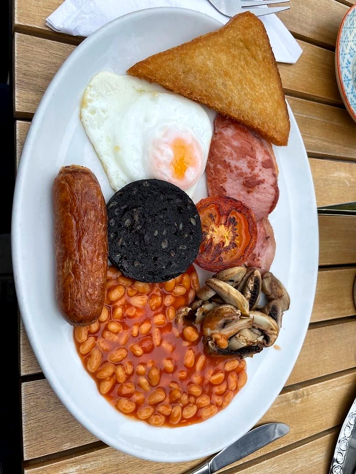 A cooked breakfast on a white oval plate: fried egg, bacon, sausage, black pudding, fried bread, baked beans, tomato and mushrooms