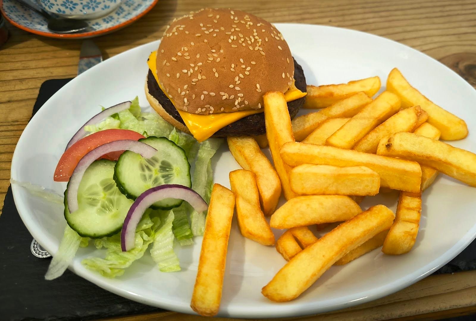 A cheeseburger in a sesame bun with chips and a side salad of lettuce, tomato, cucumber and red onion
